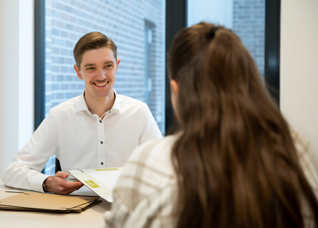 Zwei Personen sitzen sich an einem Tisch gegenüber, eine hält Dokumente in der Hand, im Hintergrund Fenster und helle Wand.