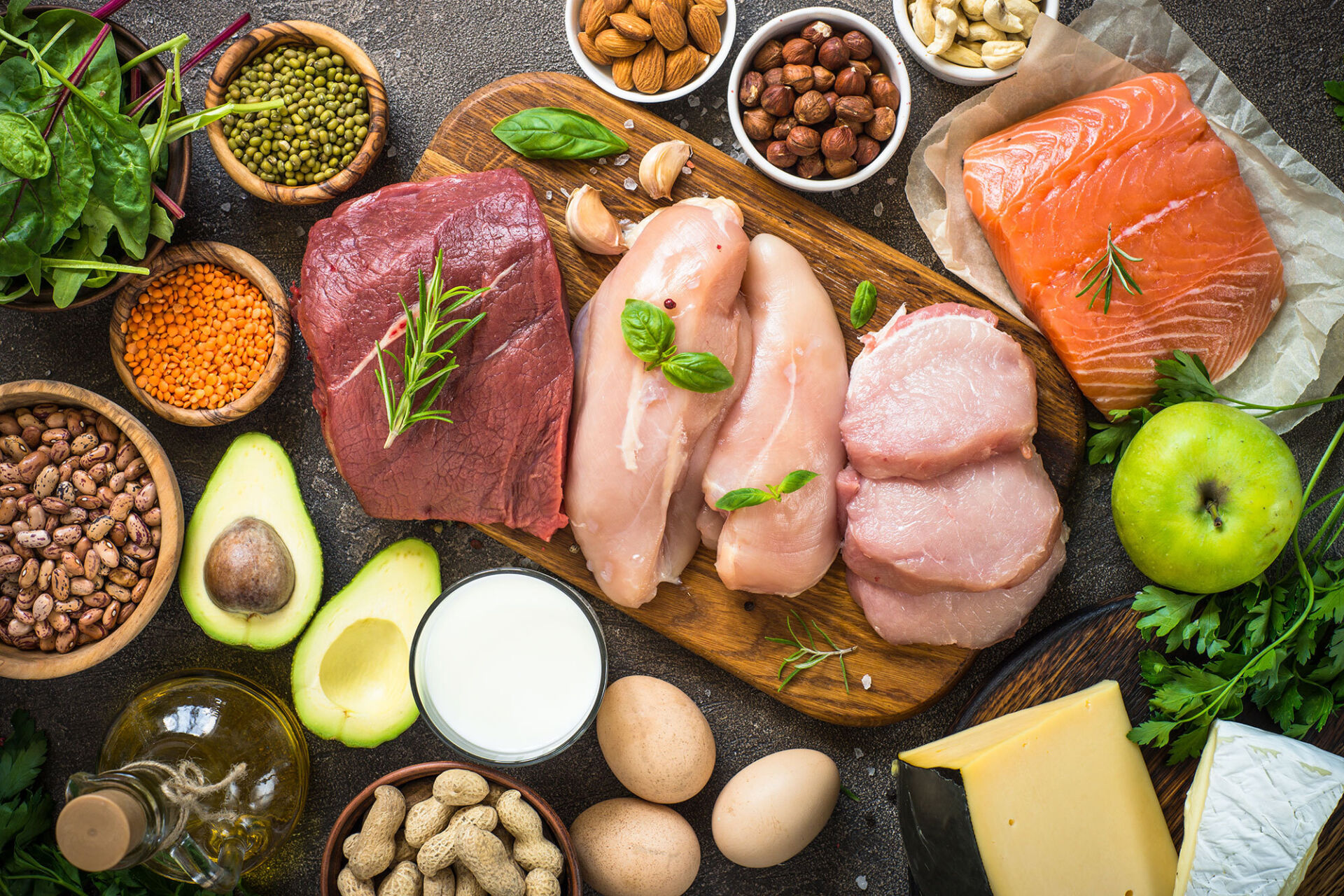 Verschiedene proteinreiche Lebensmittel wie rohes Rindfleisch, Hähnchenbrust, Lachsfilet, Eier, Käse, Nüsse, Hülsenfrüchte, Avocado, grüner Apfel und frische Kräuter auf dunklem Untergrund.
