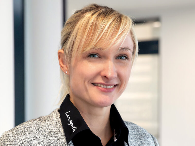 Frau mit blondem Haar, trägt grauen Blazer und schwarzes Hemd mit weißem Schriftzug 'Landguth' am Kragen.