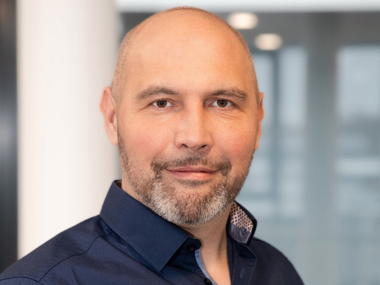 Mann mit Glatze trägt ein dunkelblaues Hemd, steht vor einem hellen, modernen Innenraum.